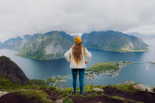 Female with long hair in yellow hat getting to the top of the mountain and enjoying a view of the ocean and the mountain peaks on Lofoten, Northern Norway