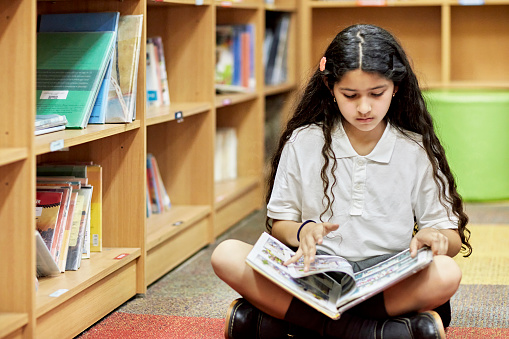 Front view of 10 year old student in uniform sitting cross-legged on floor and paging through book during class visit to library.