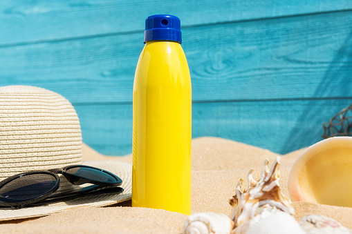 A yellow aerosol can sunscreen sitting in the sand of a beach with a straw sun hat and black sunglasses, with a turquoise blue weather plank wall and a fishing net hanging off in the background