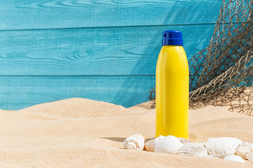 A yellow aerosol can sunscreen sitting in the sand of a beach with a turquoise blue weather plank wall with a fishing net hanging off behind and seashells in the foreground.