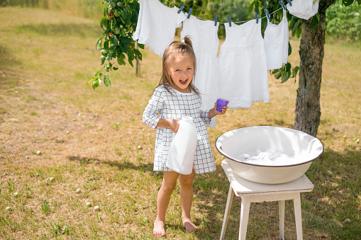 Kid is happy to help mother with household chores. The girl holds an open bottle of liquid detergent in hand. Snow-white linen dries on rope in garden under tree, developing in wind. hand wash