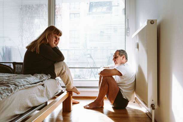 Midlife crisis, family breakdown, quarrel or divorce middle-aged married couple is sitting in bedroom after quarrel. man sits on floor with legs crossed, woman sits on bed with hands behind head arguing couple divorce family stock pictures, royalty-free photos & images