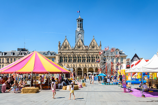 Saint-Quentin, France - August 9, 2022: Medieval facade of flamboyant gothic style of the town hall with the stands of the Circus Parc summer entertainment in the foreground on a sunny day.