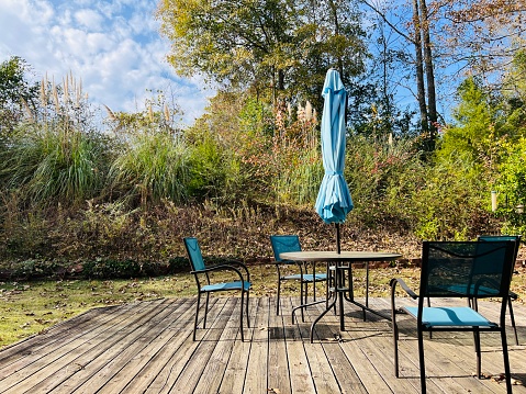 Iron patio furniture table, chairs and umbrella on a sunny day outdoors. The fabric is a sky blue, the umbrella is closed and the set is on a wooden deck in a small backyard. Surrounding by grass, trees and somewhat private.