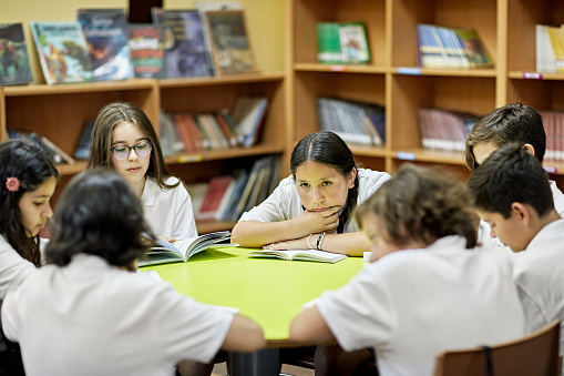 Waist-up view of elementary-age boys and girls in uniforms sitting at round table and reading their books.