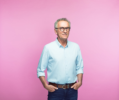 Portrait of confident retired senior man standing with hands in pockets over pink background