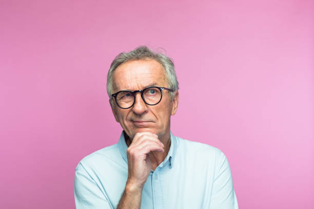 Confused retired man with hand on chin looking away Confused retired elderly man with hand on chin looking away against pink background uncertainty stock pictures, royalty-free photos & images