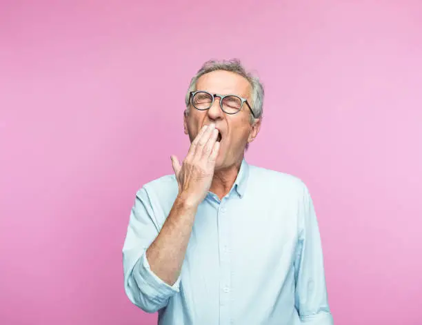 Tired senior man yawning with eyes closed against pink background