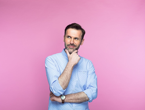 Young attractive and cheerful man of mind touching chin and deciding background isolated on pink color