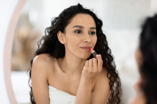 Glad pretty islamic millennial curly female in towel applying lipstick on lips, looking in mirror in bedroom interior, close up, copy space. Nude makeup at home alone, beauty care and daily procedure