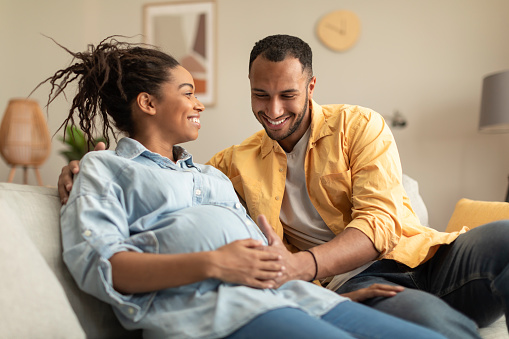 Happy african american couple expecting baby, pregnant black spouses embracing on couch at home. Smiling husband and wife resting in living room