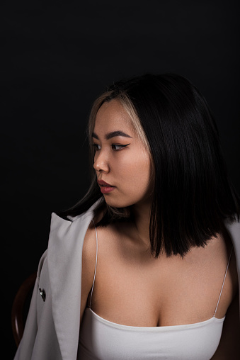Portrait of a young and attractive Asian brunette girl in stylish casual clothes in a gray jacket sitting on a chair isolated on a black studio background.