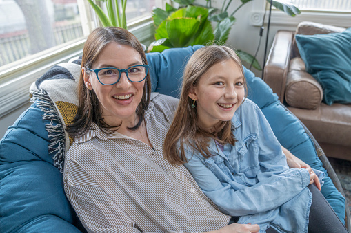 A Mother sits snuggled up in a chair with her daughter as they spend some quality time together at home.  They are both dressed casually and are smiling as they talk together.