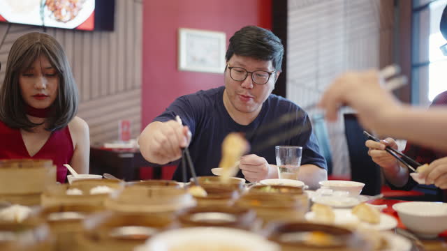 Cheerful Asian man eating a large piece of dim sum and talking with his friend in a Chinese restaurant