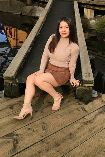 A Mexican model sitting on a ramp to a floating dock. She is wearing long black straight hair, makeup, a beige sweater and shoes with a brown mini skirt.