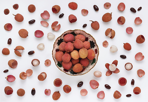 Lychee fruit in a bowl on a white background with scattered whole and peeled fruits, with seeds and peels, table top view. Decorative lychee background in flat lay style. Asian fruits also called Chinese strawberries.