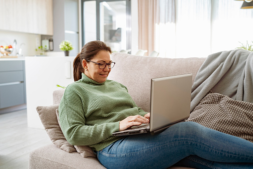 Side view of relaxed mature woman sitting on sofa using laptop for working, surfing the net, looking for online information, online shopping, etc. High resolution 42Mp indoors digital capture taken with SONY A7rII and Zeiss Batis 40mm F2.0 CF lens