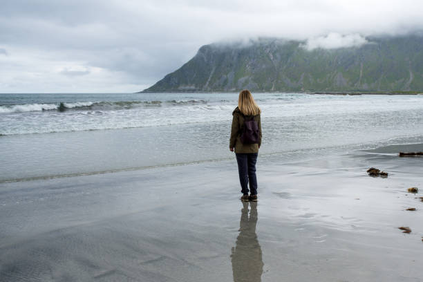 istock Young woman with a backpack walks along an empty sandy ocean beach. Town and mountains. Beautiful nature landscape. Lonely girl. Wanderlust. Travel, adventure, lifestyle. Explore North, Norway 1463534019