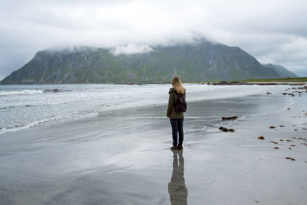 istock Young woman with a backpack walks along an empty sandy ocean beach. Town and mountains. Beautiful nature landscape. Lonely girl. Wanderlust. Travel, adventure, lifestyle. Explore North, Norway 1463534548