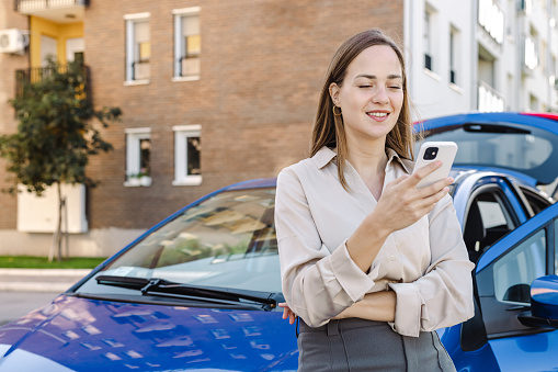 Beautiful elegant young woman leaning against her car and using a smart phone