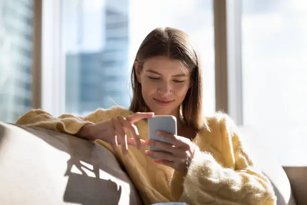 Happy peaceful smartphone user girl chatting online, using online app on mobile phone for internet communication, resting, sitting on comfortable home sofa, enjoying leisure