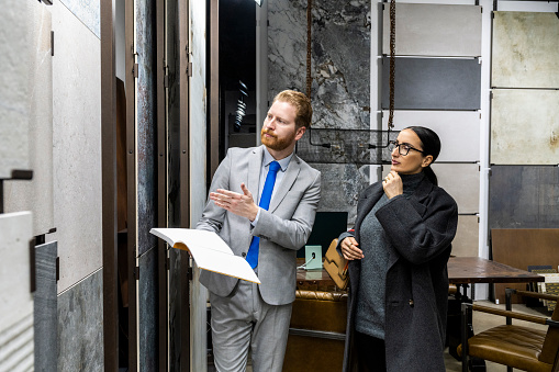 Salesman assisting an interested woman with eyeglasses wearing grey sweater and coat in a ceramics and bathroom equipment shop while choosing perfect tiles for her new home.