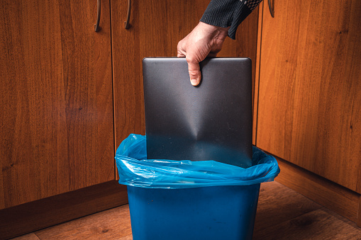 A man throws a laptop in the trash. Man's hand with a laptop and a trash can in blue..