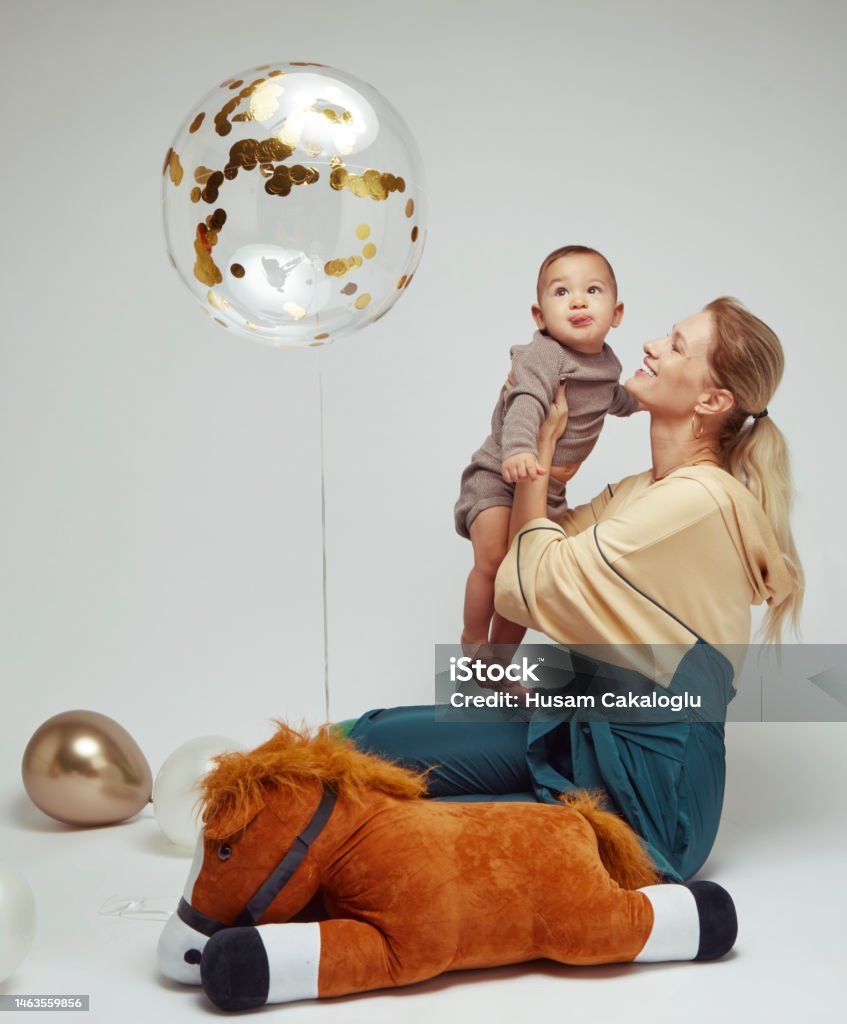 Baby boy in mother's lap looking at her flying balloon in front of white background. Balloon Stock Photo