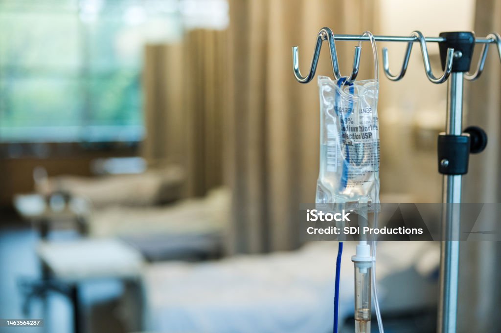 Focus on IV bag and stand in empty hospital ward The focus of the photo is on the IV bag and stand in the foreground with empty hospital beds in a row in the background. IV Drip Stock Photo