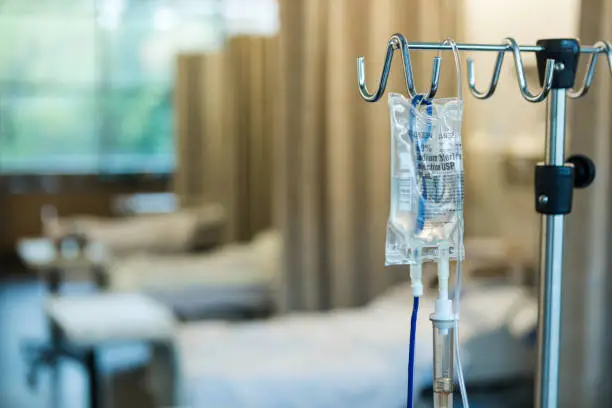 The focus of the photo is on the IV bag and stand in the foreground with empty hospital beds in a row in the background.