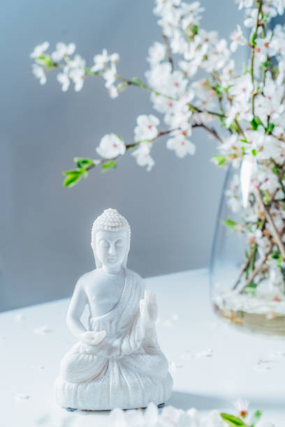 istock Decorative white Buddha statuette with blooming tree branches in the vase on the light background. Meditation and relaxation ritual. Buddha birthday. Vertical card. Selective focus 1463568187