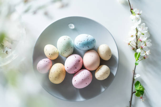 istock Top view colored easter eggs on the gray plate, blooming tree branch in vase and flower petals on the white table. Happy Easter and springtime card. Minimalism and simplicity. Selective focus. 1463568212