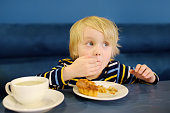 Cute blonde boy eating pie and drinking tea in cafe or restaurant. Child eating with his hands. Healthy meal for kids.