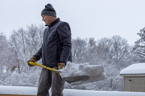 This image shows a close-up partial view of an adult man with a shovel full of snow on a wooden deck on an overcast winter day.