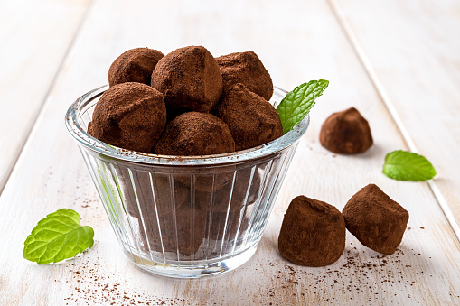 Chocolate truffles in a glass bowl closeup. Delicious dark chocolate candies and green mint leaves over white wooden table. Homemade dessert, gourmet confection recipe. Sweet food concept. Front view.