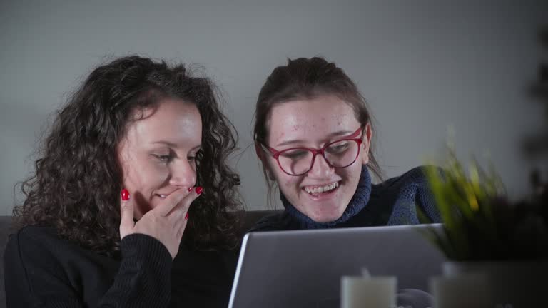 Two young beautiful female hiccup and smiling student with laptop sitting on sofa in an appartment