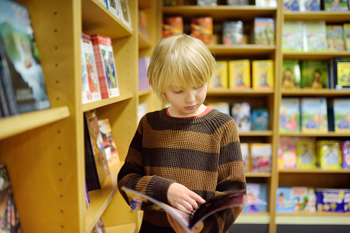 A preteen boy leafing through a book while standing at the bookshelfs in a school library or bookstore. Smart kid reading comics or adventure book