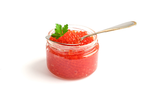 Red caviar in glass jar with spoon and green parsley leaf on top. Isolated on white background. Seafood. Delicatessen, gourmet food. Russian national cuisine.