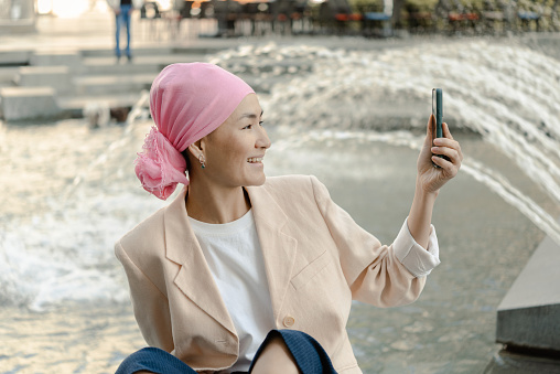 Portrait of an Asian female cancer patient with a headscarf taking a break in the city and making a video call
