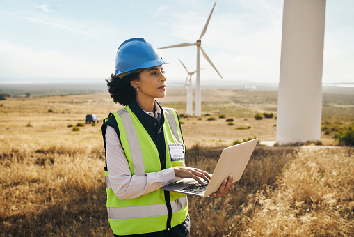 Windmill on farm, black woman in clean energy typing on laptop and electricity generation with wind turbines in Africa. Renewable power, female engineer in agriculture environment and safety gear