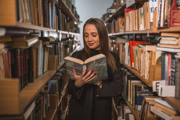 istock Beautiful librarian is reading a book while standing in the library between the bookshelves 1463637347