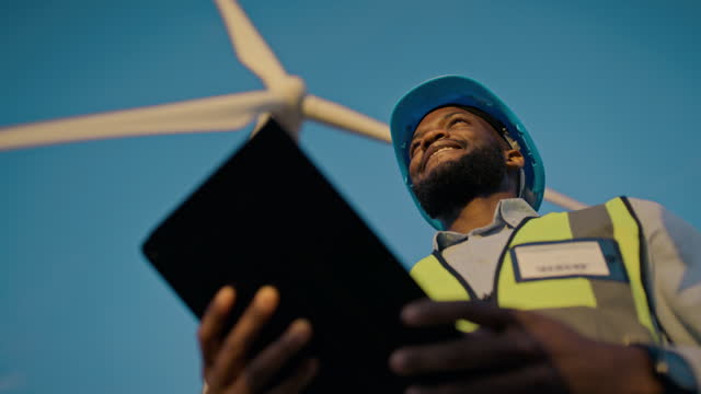 Low angle of black man, wind turbine and tablet app for renewable energy, construction and industrial architecture. Contractor, engineering and digital technology on blue sky of future sustainability