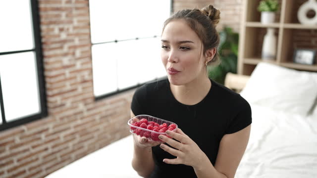 Young beautiful hispanic woman eating raspberries sitting on bed at bedroom