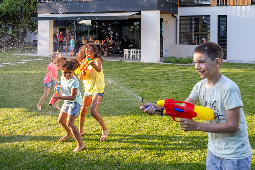 Group of young kids having water fight against house with squirt guns in backyard.