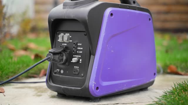 A small gasoline inverter generator with a wire connected to a socket works on the street close-up. Purple power generator with a plastic case. Alternative power source in case of blackout