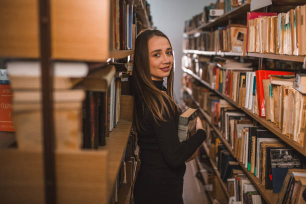 istock Side view of female librarian holding a book while standing leaning against bookshelf in library and looking at camera 1463662973