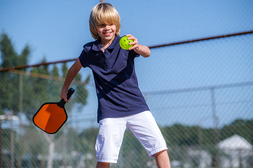 Happy blonde boy playing pickleball game, hitting pickleball yellow ball with paddle, outdoor sport leisure kids activity
