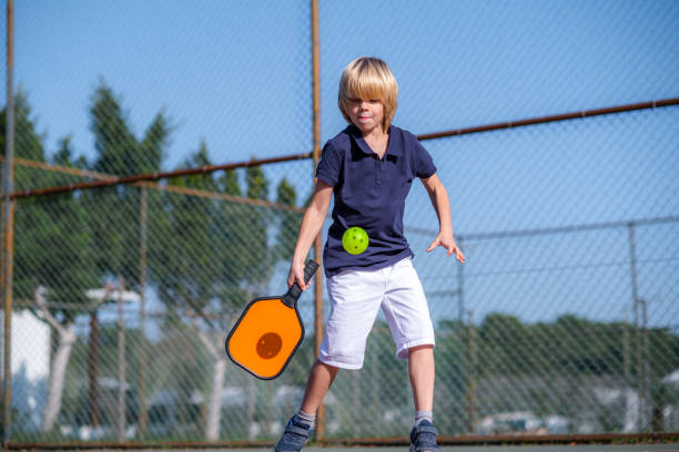 Happy blonde boy playing pickleball game, hitting pickleball yellow ball with paddle, outdoor sport leisure kids activity. stock photo