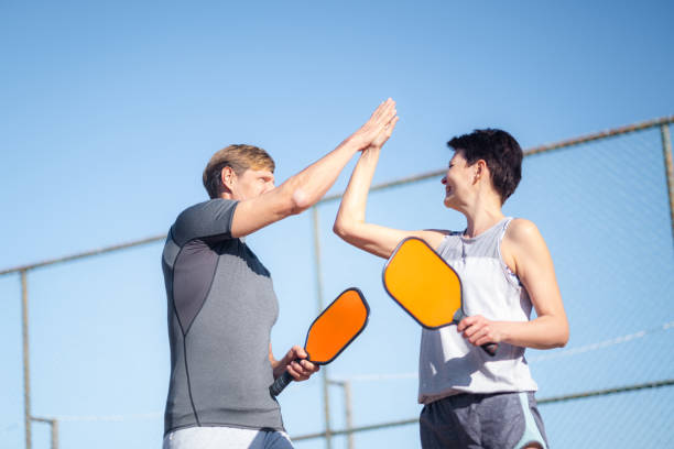 Laughing couple playing pickleball game, hitting pickleball yellow ball with paddle, outdoor sport leisure activity, celebrating victory . stock photo