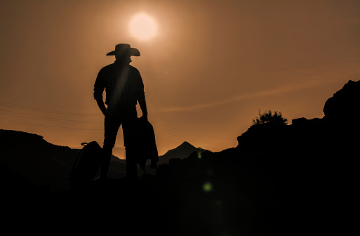 Silhouette of adult man in cowboy hat against mountain and sky during sunset